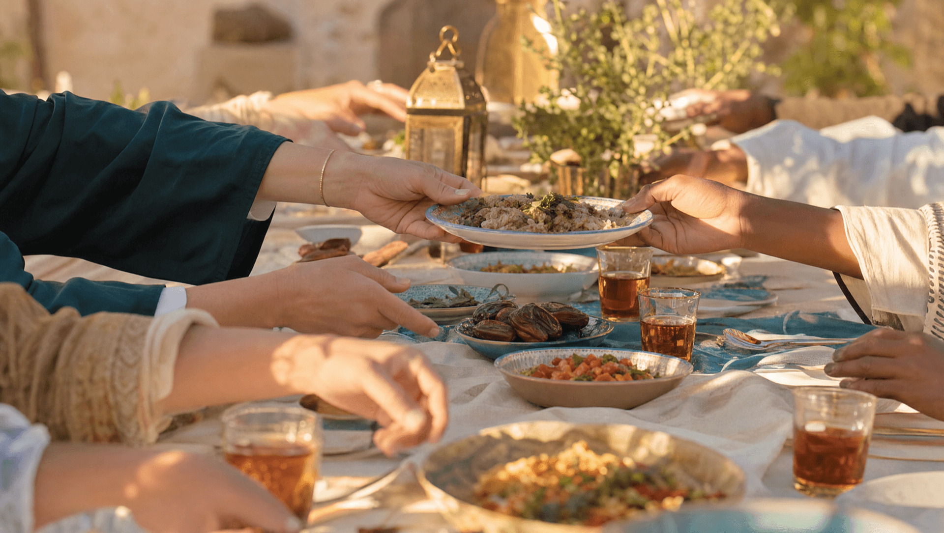 A shared meal at a long outdoor table in golden light, hands of many people passing dishes of food.